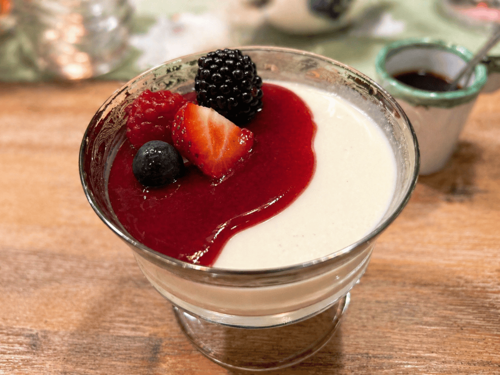 Panna Cotta angerichtet in einem altmodischen Dessertglas mit Beerenspiegel und Waldfrüchten auf einem rustikalen Holztisch