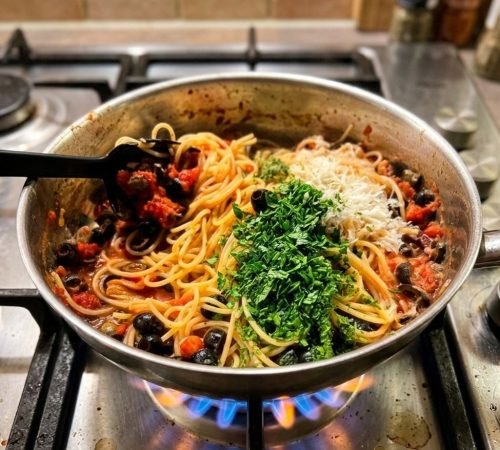 Pfanne auf einem Gasherd befüllt mit Spaghetti, Parmesan, Tomatensauce, Oliven und frischer Petersilie